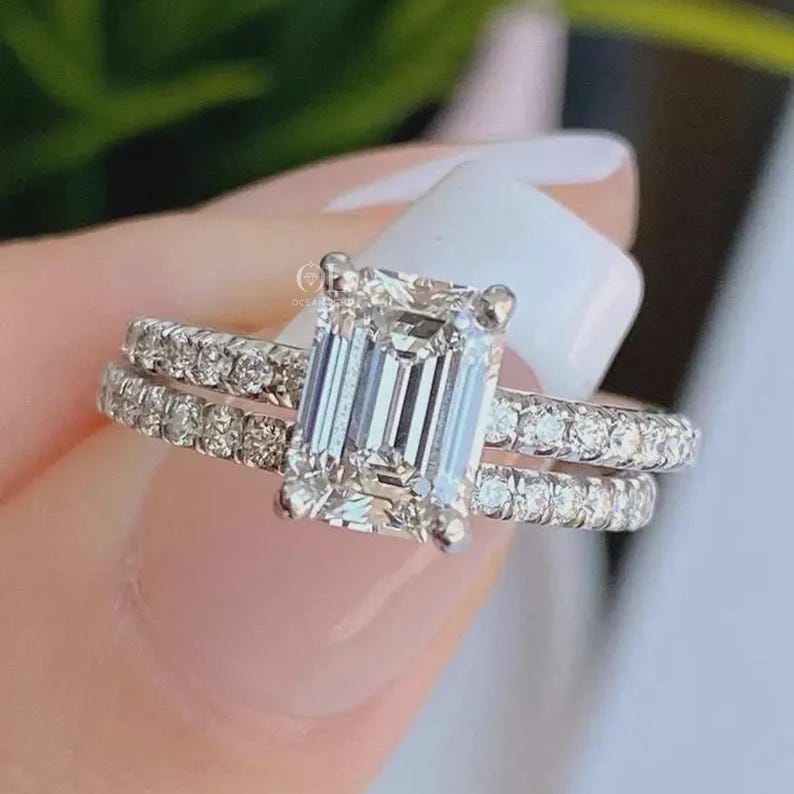 May include: A silver-toned ring featuring a large emerald-cut center stone held by four prongs, flanked by a band encrusted with small round pav&eacute;-set stones. The metal band displays a bright, reflective finish with clear, faceted gemstones.