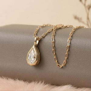 May include: A gold necklace featuring a pear-shaped pendant with a clear, faceted stone set in a gold frame. The pendant hangs from a delicate gold chain with elongated oval links. The metal has a polished finish and a warm, yellow gold tone.