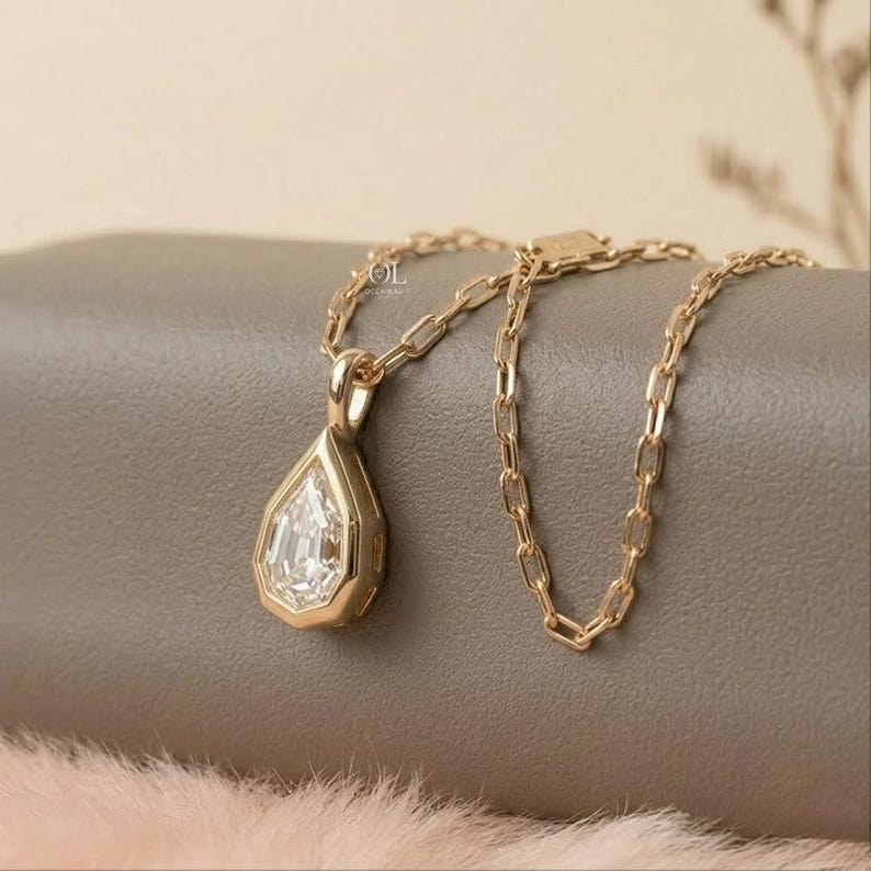 May include: A gold necklace featuring a pear-shaped pendant with a clear, faceted stone set in a gold frame. The pendant hangs from a delicate gold chain with elongated oval links. The metal has a polished finish and a warm, yellow gold tone.