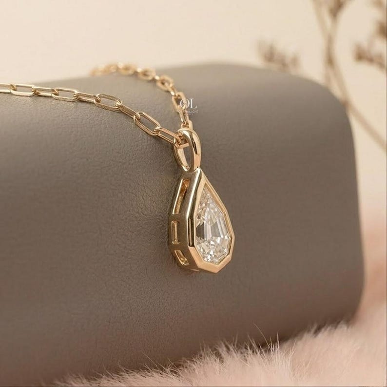 May include: A gold pendant necklace featuring a pear-shaped faceted clear gemstone set in a polished gold frame. The pendant hangs from a delicate gold chain with elongated rectangular links. The metal has a warm, reflective, metallic finish.