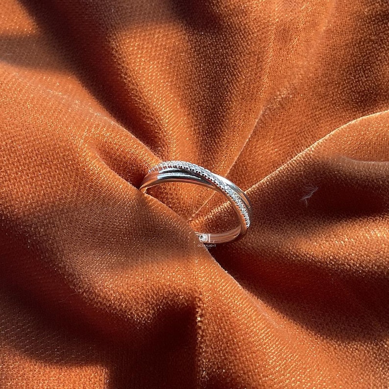 Puede incluir: Un anillo plateado con un dise&ntilde;o entrecruzado, engastado con peque&ntilde;as piedras preciosas transparentes. El anillo se exhibe sobre una tela texturizada de color &oacute;xido, creando un contraste entre el metal brillante y el fondo mate. El dise&ntilde;o del anillo es elegante y moderno.