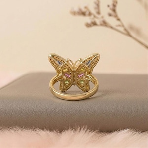 May include: A gold-toned ring featuring a butterfly-shaped centerpiece. The butterfly wings are decorated with small, rectangular, multi-colored gemstones in shades of pink, yellow, and green, surrounded by tiny clear stones along the metal frame.
