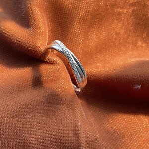 Puede incluir: Un anillo plateado con un dise&ntilde;o entrecruzado, engastado con peque&ntilde;as piedras brillantes. El anillo se exhibe sobre una tela texturizada de color &oacute;xido, resaltando su metal pulido y el brillo de las piedras. El dise&ntilde;o del anillo es moderno y elegante.