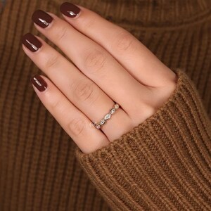 May include: A gold ring with a series of small, clear gemstones set in a repeating pattern. The ring is worn on a finger, and the hand is resting on a brown sweater.