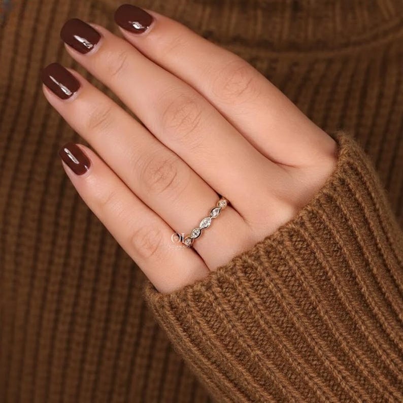 May include: A gold ring with a series of small, clear gemstones set in a repeating pattern. The ring is worn on a finger, and the hand is resting on a brown sweater.
