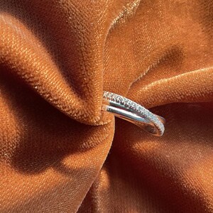 Puede incluir: Un anillo de plata con un dise&ntilde;o de doble banda, una banda est&aacute; incrustada con peque&ntilde;as piedras preciosas transparentes. El anillo se coloca sobre un fondo de tela texturizada de color &oacute;xido, creando un contraste de texturas y colores.