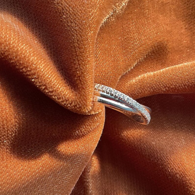 Puede incluir: Un anillo de plata con un dise&ntilde;o de doble banda, una banda est&aacute; incrustada con peque&ntilde;as piedras preciosas transparentes. El anillo se coloca sobre un fondo de tela texturizada de color &oacute;xido, creando un contraste de texturas y colores.