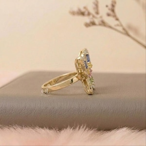 May include: A gold ring featuring a butterfly-shaped centerpiece adorned with small, multi-colored baguette-cut gemstones including blue, yellow, pink, and green stones, with a smooth, polished gold band and a small logo mark on the side.