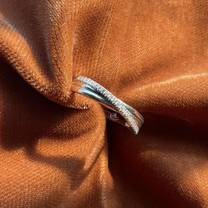 Puede incluir: Un anillo plateado con un dise&ntilde;o entrecruzado, adornado con peque&ntilde;as piedras brillantes a lo largo de una banda. El anillo se encuentra sobre un fondo de tela de terciopelo marr&oacute;n suave, creando un contraste de texturas.