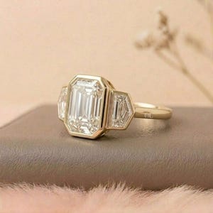 Puede incluir: Un anillo de oro con una gran gema clara rectangular en el centro, flanqueada por dos gemas m&aacute;s peque&ntilde;as en forma de trapecio. El anillo tiene una banda simple y est&aacute; sobre una superficie neutra.