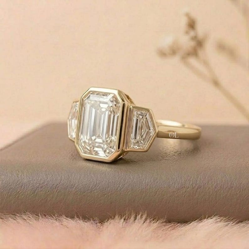 Puede incluir: Un anillo de oro con una gran gema clara rectangular en el centro, flanqueada por dos gemas m&aacute;s peque&ntilde;as en forma de trapecio. El anillo tiene una banda simple y est&aacute; sobre una superficie neutra.