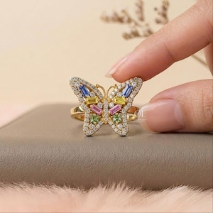 May include: A gold ring featuring a butterfly-shaped centerpiece adorned with small clear pav&eacute; stones and larger baguette-cut stones in blue, yellow, pink, and green. The butterfly design is symmetrical, with the stones set in a gold metal frame.