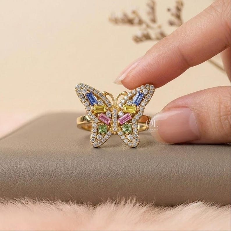 May include: A gold ring featuring a butterfly-shaped centerpiece adorned with small clear pav&eacute; stones and larger baguette-cut stones in blue, yellow, pink, and green. The butterfly design is symmetrical, with the stones set in a gold metal frame.