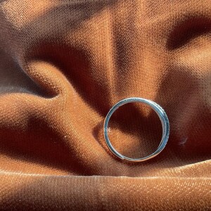 Puede incluir: Un anillo plateado con una banda de peque&ntilde;as piedras transparentes. El anillo se encuentra sobre un fondo de tela marr&oacute;n aterciopelada. El dise&ntilde;o del anillo es sencillo y elegante.