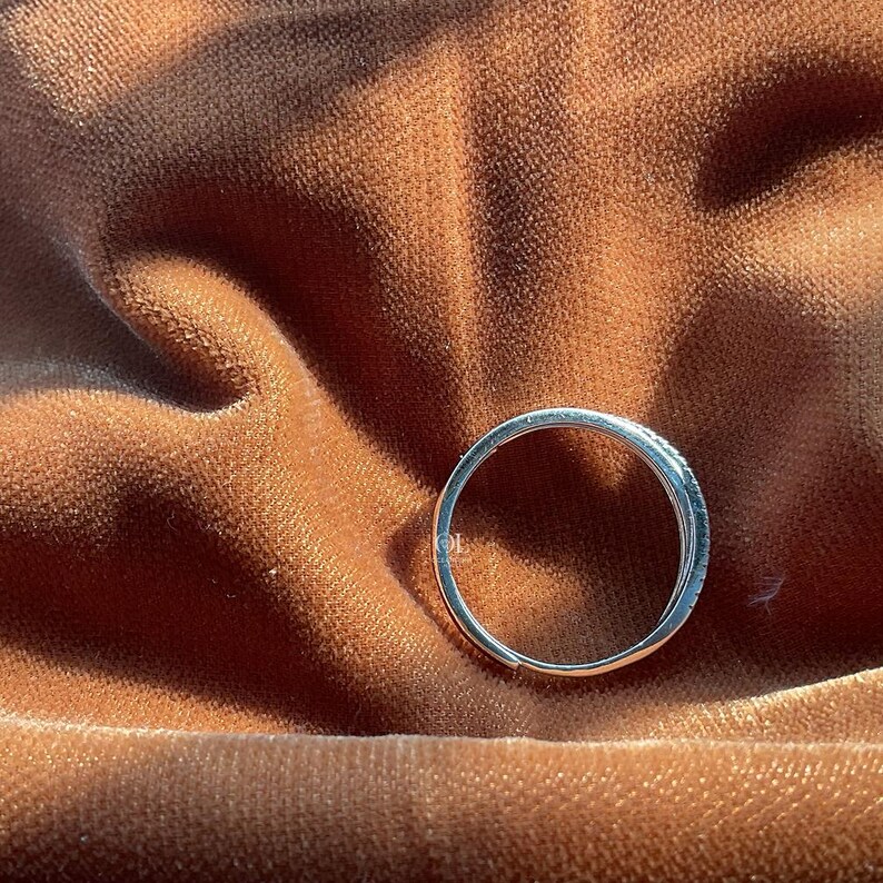 Puede incluir: Un anillo plateado con una banda de peque&ntilde;as piedras transparentes. El anillo se encuentra sobre un fondo de tela marr&oacute;n aterciopelada. El dise&ntilde;o del anillo es sencillo y elegante.