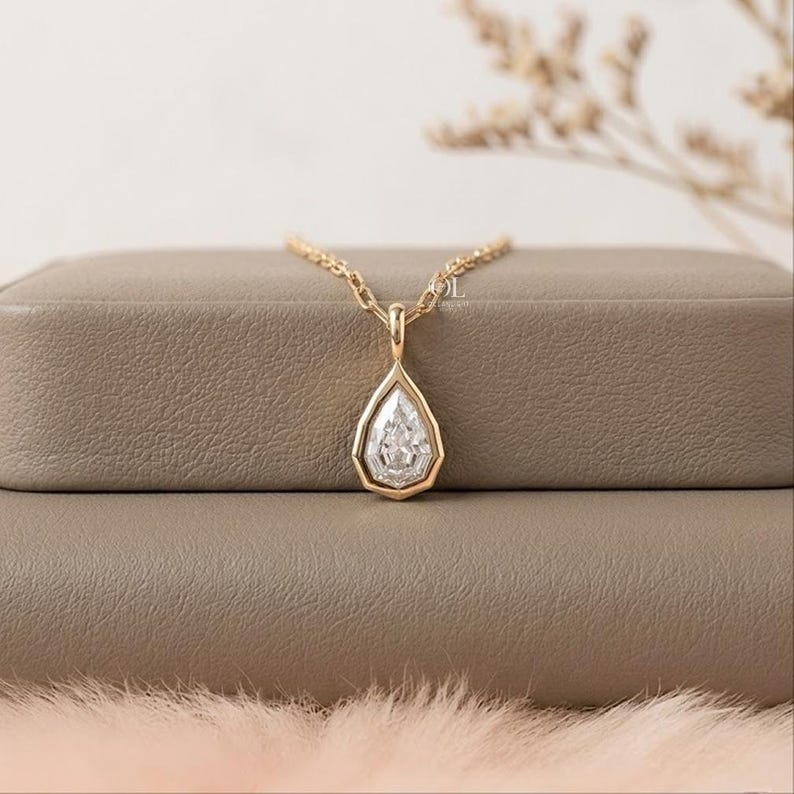 May include: A gold necklace featuring a pear-shaped pendant with a central clear stone surrounded by a gold bezel setting. The pendant hangs from a delicate gold chain, showcasing a faceted design that reflects light across its surface.