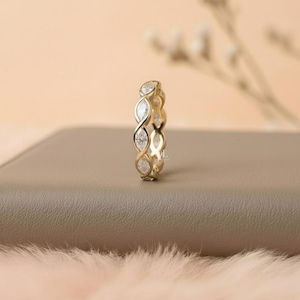 May include: A gold-colored ring with a continuous design of marquise-cut gemstones. The ring is set on a neutral-toned surface, with a soft, blurred background. The ring's design features a repeating pattern of intertwined shapes.