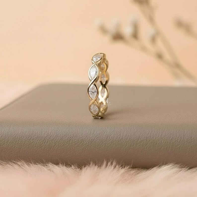 May include: A gold-colored ring with a continuous design of marquise-cut gemstones. The ring is set on a neutral-toned surface, with a soft, blurred background. The ring's design features a repeating pattern of intertwined shapes.
