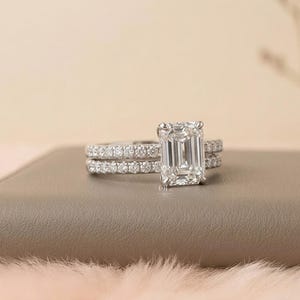 May include: A diamond engagement ring with an emerald cut center stone and a matching band. The rings are set in a silver metal and are displayed on a neutral surface. The center stone is clear and sparkling, and the band is encrusted with smaller diamonds.