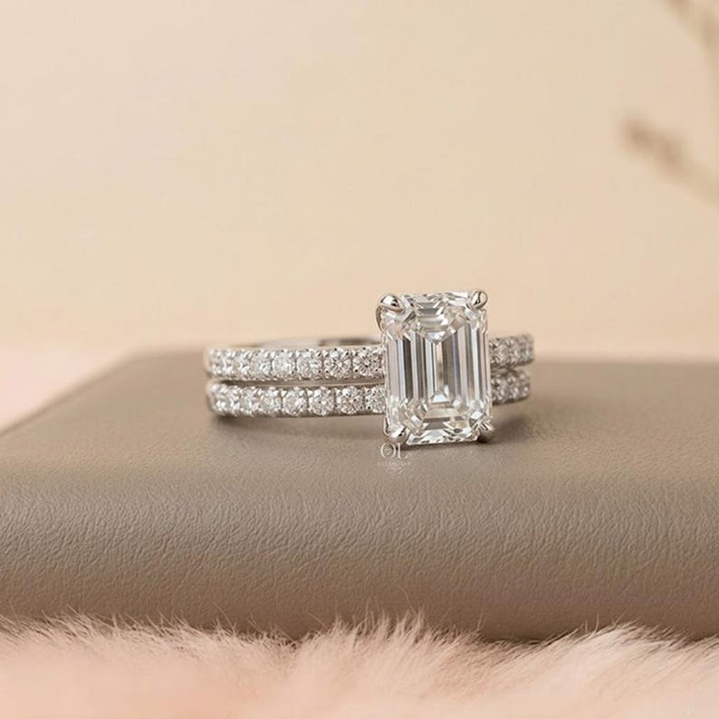 May include: A diamond engagement ring with an emerald cut center stone and a matching band. The rings are set in a silver metal and are displayed on a neutral surface. The center stone is clear and sparkling, and the band is encrusted with smaller diamonds.