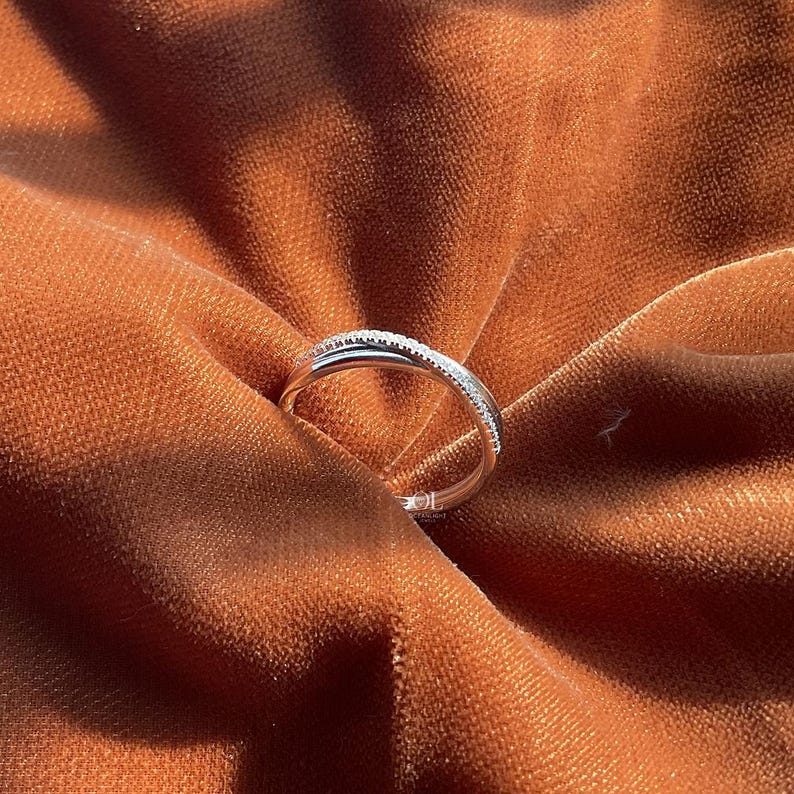 Puede incluir: Un anillo de plata con un dise&ntilde;o delicado e entrelazado. Una de las bandas est&aacute; adornada con peque&ntilde;as piedras brillantes, creando un detalle sutil pero llamativo. El anillo descansa sobre una tela texturizada de color &oacute;xido.