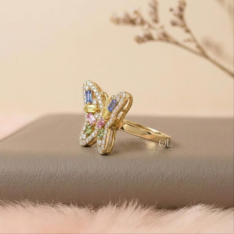 May include: A gold ring featuring a butterfly-shaped centerpiece adorned with small clear stones and baguette-cut stones in blue, yellow, pink, and green. The ring band is smooth and polished gold, with the letters OL visible on the side of the setting.