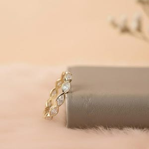May include: A gold-colored ring with a continuous pattern of clear, oval-shaped stones. The ring is set against a soft, peach-colored background with a neutral-colored block. The ring's design features a repeating, intertwined pattern.