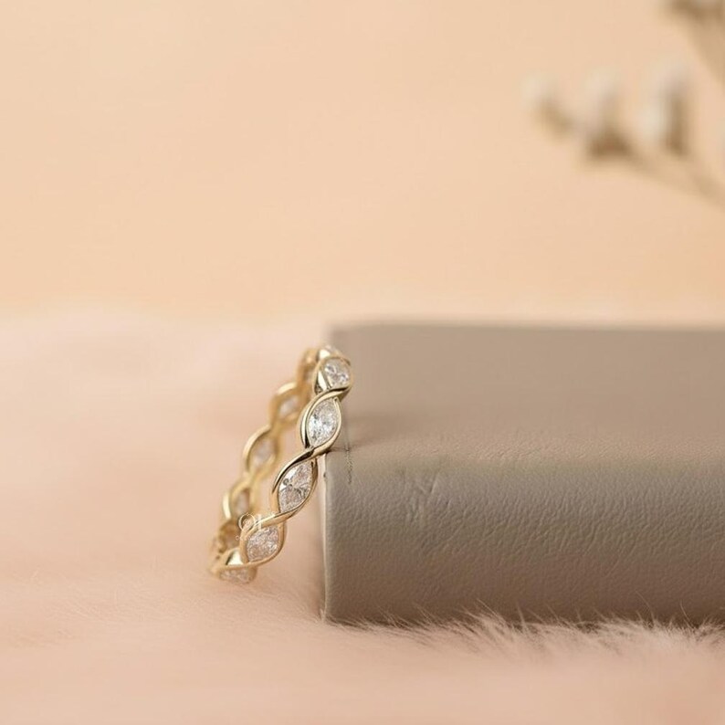 May include: A gold-colored ring with a continuous pattern of clear, oval-shaped stones. The ring is set against a soft, peach-colored background with a neutral-colored block. The ring's design features a repeating, intertwined pattern.