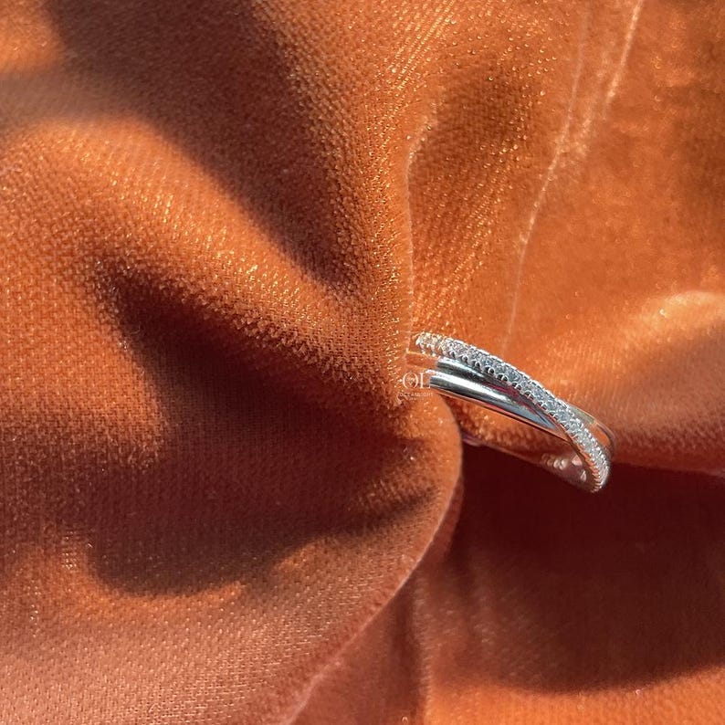 Puede incluir: Un anillo plateado con un dise&ntilde;o entrecruzado, adornado con peque&ntilde;as piedras brillantes. El anillo se encuentra sobre un fondo de tela de terciopelo suave de color marr&oacute;n anaranjado, creando un contraste de texturas y colores. El dise&ntilde;o del anillo es moderno y elegante.