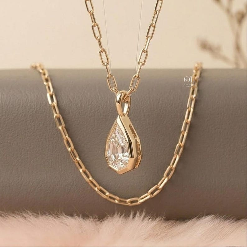 May include: A gold-toned necklace featuring a teardrop-shaped pendant with a clear, faceted stone set in a metallic frame. The pendant hangs from a delicate chain composed of elongated, rectangular links, creating a structured and refined appearance.