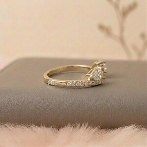 May include: A gold-colored ring with two pear-shaped gemstones and small, round accent stones. The ring has a delicate band and a unique design, perfect for a special occasion. The ring is set on a neutral surface.