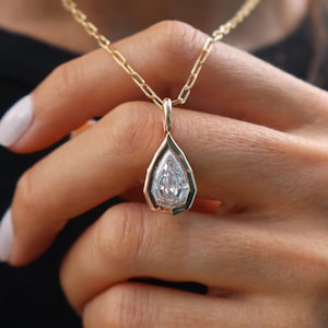 May include: A gold necklace featuring a pear-shaped clear gemstone pendant set in a geometric gold bezel. The pendant hangs from a delicate gold chain with elongated rectangular links, showcasing a polished metallic finish and a faceted stone.