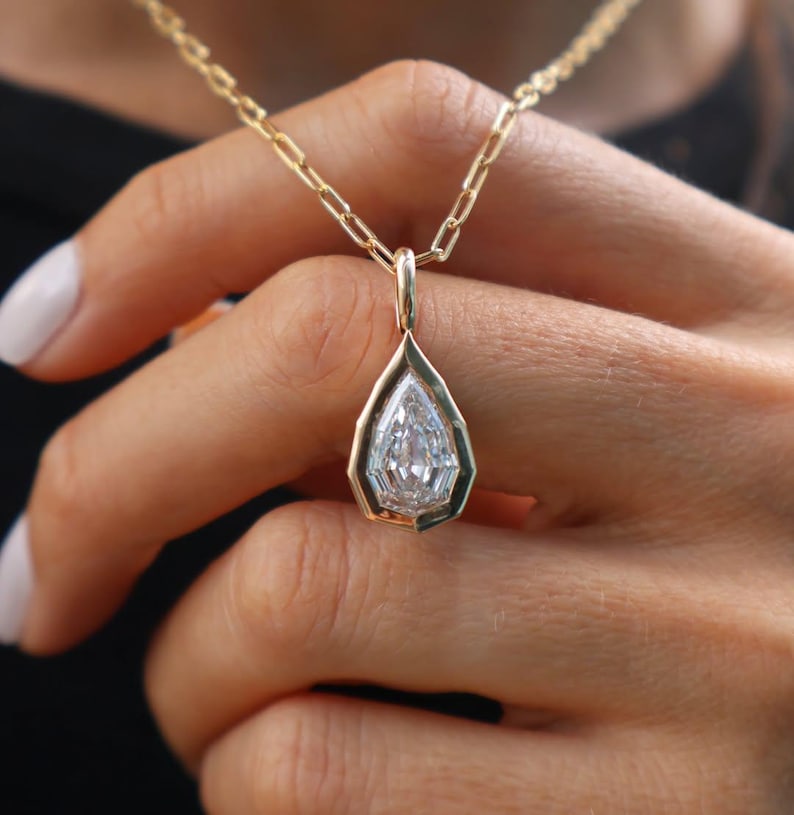 May include: A gold necklace featuring a pear-shaped clear gemstone pendant set in a geometric gold bezel. The pendant hangs from a delicate gold chain with elongated rectangular links, showcasing a polished metallic finish and a faceted stone.
