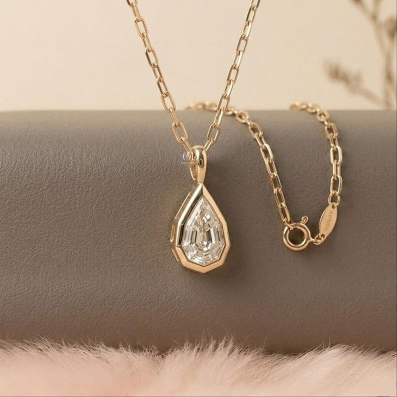 May include: A gold necklace featuring a pear-shaped pendant with a faceted clear stone set in a gold bezel. The pendant hangs from a delicate gold chain with rectangular links. A small oval tag is attached to the chain near the circular clasp.