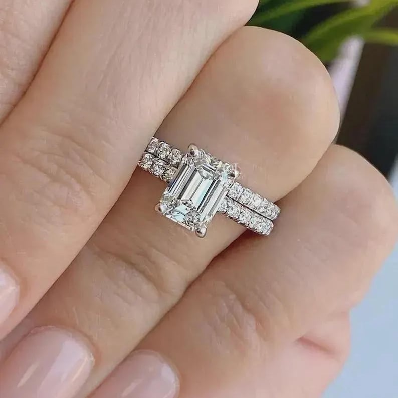 May include: A diamond engagement ring set featuring an emerald-cut center stone and a pav&eacute; band. The ring is set on a finger, showcasing its brilliance. The band is adorned with smaller diamonds, adding to its sparkle. A matching band is also present.