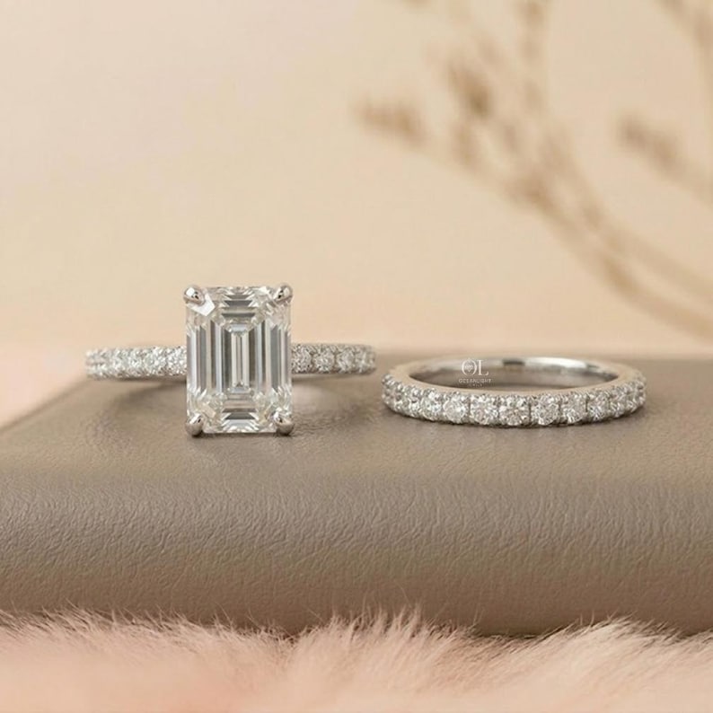 May include: A two-piece ring set featuring a large emerald-cut diamond engagement ring and a matching diamond band. The rings are set in a silver metal, with small diamonds lining the band of the engagement ring and the entire band of the wedding ring.