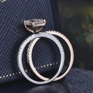 May include: A close-up of two rings. The first ring has a large, rectangular, clear gemstone set in a rose gold band. The second ring is a silver band with small, clear gemstones. The rings are on a dark blue surface.