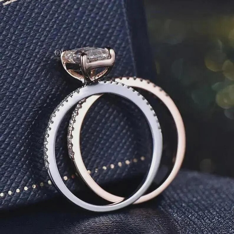 May include: A close-up of two rings. The first ring has a large, rectangular, clear gemstone set in a rose gold band. The second ring is a silver band with small, clear gemstones. The rings are on a dark blue surface.