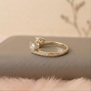 May include: A gold ring with two clear, round-cut gemstones. The band is also adorned with smaller, clear gemstones. The ring is set on a neutral-toned surface, with a soft, pinkish-white texture in the foreground.