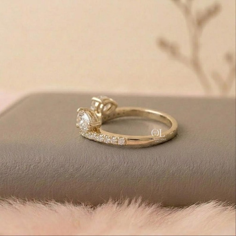 May include: A gold ring with two clear, round-cut gemstones. The band is also adorned with smaller, clear gemstones. The ring is set on a neutral-toned surface, with a soft, pinkish-white texture in the foreground.