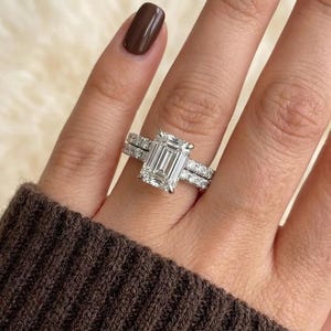 May include: A silver engagement ring with an emerald-cut diamond and a diamond band. The ring is on a finger with a brown nail polish. The ring is set against a soft, neutral background.