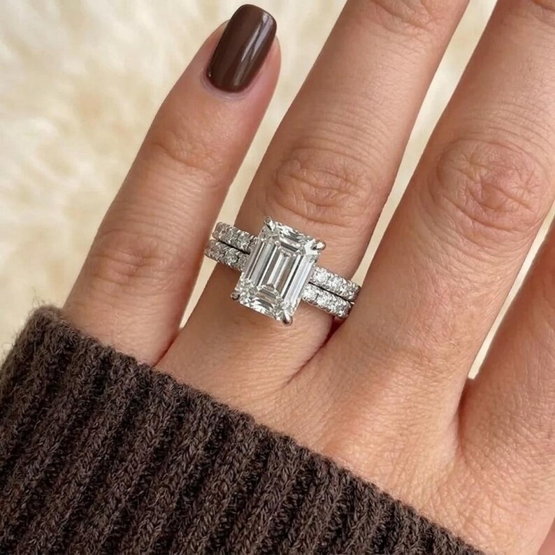 May include: A silver engagement ring with an emerald-cut diamond and a diamond band. The ring is on a finger with a brown nail polish. The ring is set against a soft, neutral background.