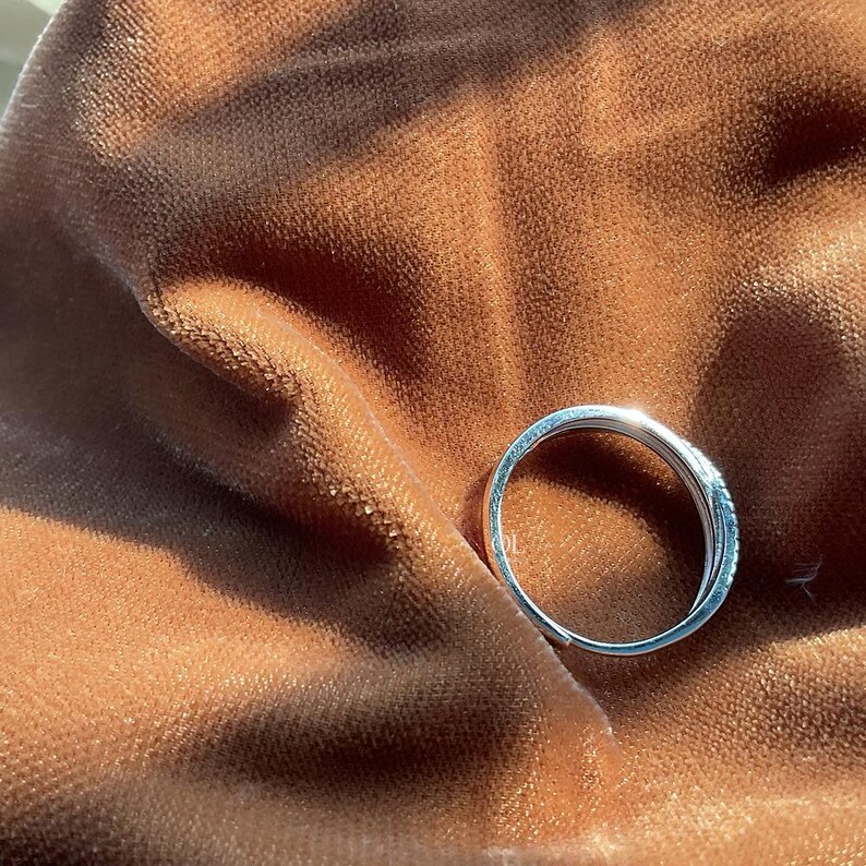 Puede incluir: Un anillo de plata con un dise&ntilde;o delicado, sobre un fondo de rico terciopelo marr&oacute;n. La superficie pulida del anillo refleja la luz, resaltando sus intrincados detalles. El anillo parece ser una banda, posiblemente para una boda o promesa.