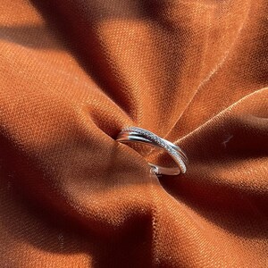 Puede incluir: Un anillo plateado con un dise&ntilde;o entrecruzado, engastado con peque&ntilde;as piedras brillantes. El anillo se muestra sobre una tela texturizada de color &oacute;xido, creando un contraste entre el metal fr&iacute;o y el fondo c&aacute;lido. El dise&ntilde;o del anillo es moderno y elegante.