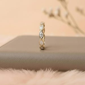 May include: A gold-colored ring with a continuous design of alternating marquise-cut and round-cut clear gemstones. The ring is set on a neutral-toned surface with a soft, blurred background.