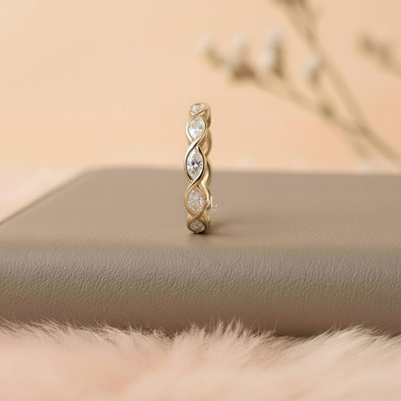 May include: A gold-colored ring with a continuous design of alternating marquise-cut and round-cut clear gemstones. The ring is set on a neutral-toned surface with a soft, blurred background.