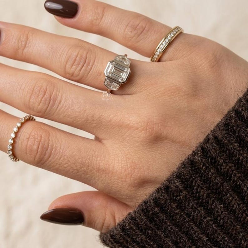 Puede incluir: Una mano con tres anillos: una banda de oro con peque&ntilde;os diamantes, una banda de oro con una fila de diamantes y un anillo de plata con una piedra central de talla esmeralda y piedras laterales. La mano tiene esmalte de u&ntilde;as marr&oacute;n oscuro y lleva un su&eacute;ter marr&oacute;n.