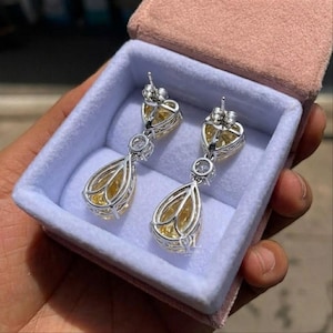 May include: A pair of elegant drop earrings with a heart-shaped top and a teardrop-shaped bottom. The earrings feature a yellow gemstone and are set in a silver-colored metal. They are displayed in a pink jewelry box with a light blue interior.
