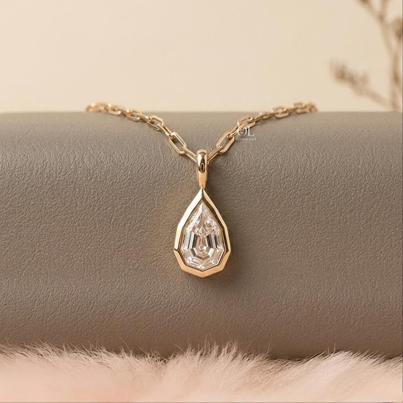 May include: A gold necklace featuring a pear-shaped pendant with a faceted clear stone set in a gold bezel. The pendant hangs from a gold chain with elongated rectangular links. The metal has a warm yellow tone and a polished finish.