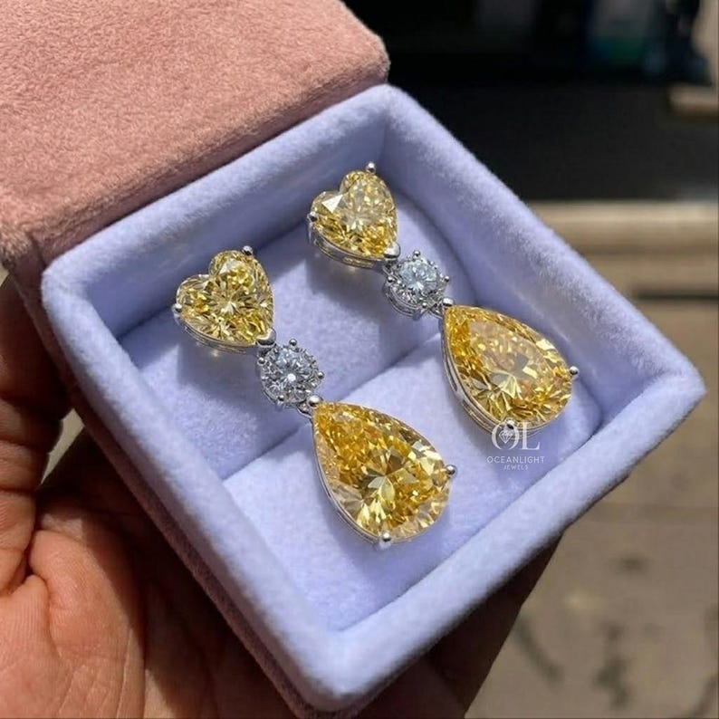 May include: A pair of elegant earrings with a heart-shaped top and a pear-shaped bottom, both in a vibrant yellow hue. The earrings feature a small, round, clear stone between the two yellow stones. The earrings are displayed in a pink and blue jewelry box.